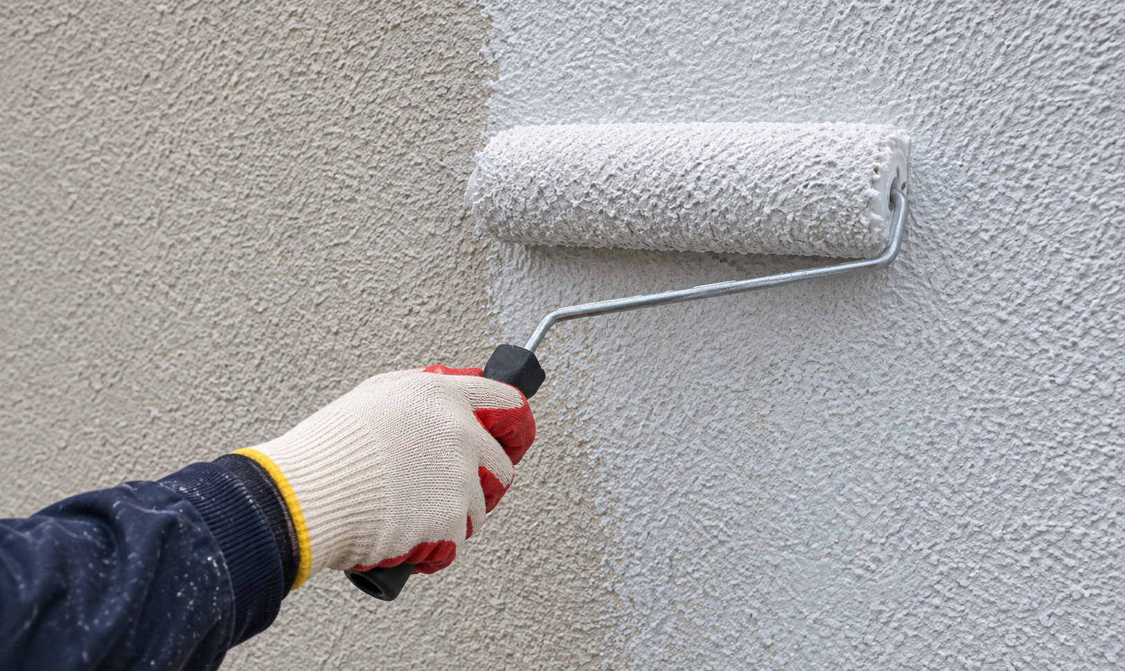 Red-coated work glove holding a roller applying exterior wall coating